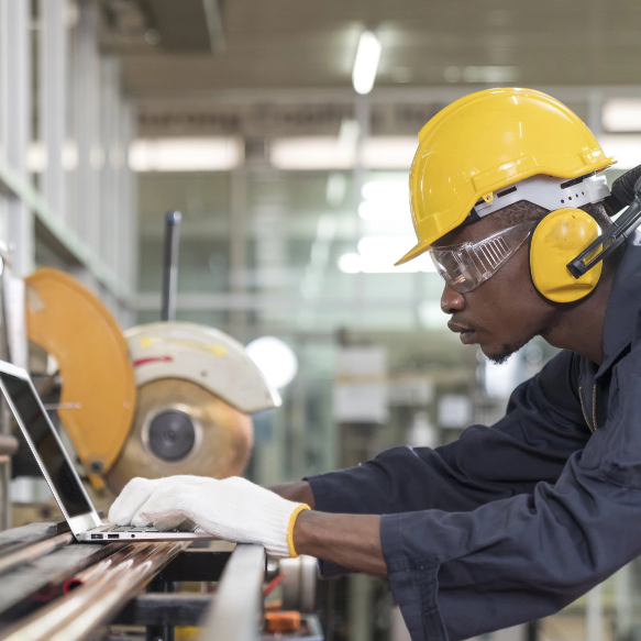 A worker operates a laptop in a warehouse while wearing personal protective equipment